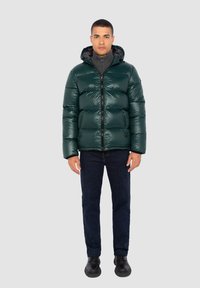 Chaqueta de plumas en verde oscuro, con un acabado brillante, cierre de cremallera, capucha y bolsillos laterales. Combinada con vaqueros oscuros y botas negras.