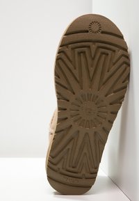 Suela de bota UGG marrón con un distintivo patrón de huellas en zigzag, colocada verticalmente contra una pared y un suelo blancos.