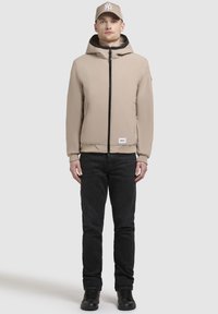 Giacca beige con zip e cappuccio, interno nero e polsini elastici. Abbinata a jeans neri e sneaker nere, indossata con un cappello beige.