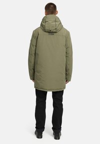 Maslinasto zeleni debeli parka kaput s kapuljačom, ravnog kroja i s teksturiranim završetkom. Ima logotip na stražnjoj strani. Nosi se preko crnih hlača.