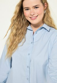 Lyseblå skjorte med knaplukning lavet af blødt stof, med klassisk krave og lange ærmer, der har en afslappet pasform og en subtil glans.