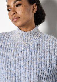Blauer und beige gestrickter Pullover mit einem hohen Rippkragen, der ein strukturiertes Muster und ein weiches, flauschiges Material aufweist.
