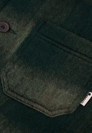 Tessuto di lana verde scuro con una morbida texture, dotato di una tasca con pattina e un bottone marrone. Piccola etichetta visibile sul bordo.
