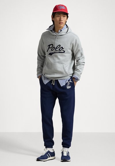 Felpa grigia con cappuccio e logo "Polo", camicia a righe blu sotto, pantaloni jogger blu navy e cappellino rosso. Sneakers con dettagli blu e bianchi.
