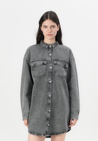 Grå denim skjortekjole med lange ermer, knappelukking, to frontlommer og rundet kant. Myk tekstur med en falmet utseende.