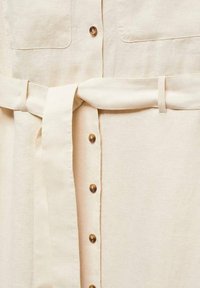 Robe en lin beige avec des boutons sur le devant, deux poches poitrine et une ceinture en tissu assortie nouée à la taille.