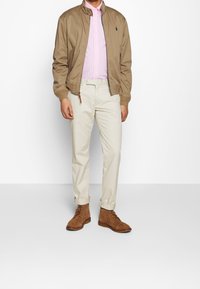 Tan Bomberjacke über einem rosa-weiß gestreiften Hemd, cremefarbige Hosen und braune Wildlederstiefel. Auffällige Nähte und Hardware-Details sichtbar.