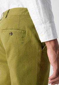 Des chinos en coton verts avec une poche arrière dotée d'un bouton et de coutures décoratives en couleurs contrastées. Le tissu a une texture lisse.