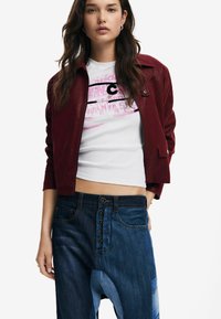 Cranberry-farbenes Kunstlederjackett mit taillierter Passform, kombiniert mit einem weißen, bedruckten Cropped-Top und weit geschnittenen blauen Jeans im Denim-Look mit Knopfdetails.