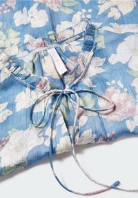 Blouse bleue à fleurs blanches et roses, col à volants et cordon à nouer au niveau de l'encolure, posée à plat sur une surface blanche.