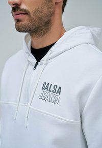 Sudadera blanca con cremallera hecha de tela suave, que presenta un logotipo negro "SALSA JEANS" en el pecho, con cordones y un diseño elegante y moderno.