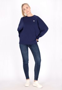 Mörkblå oversized sweatshirt med rund halsringning, ribbade ärmar och en liten vit paraplylogotyp på bröstet, kombinerad med mörka jeans och vita sneakers.
