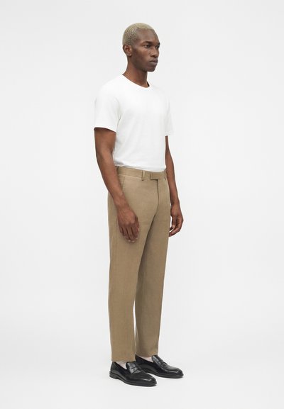 Άνδρας με κοντά ξανθά μαλλιά στέκεται, φοράει λευκό μπλουζάκι, μπεζ παντελόνι, λευκές κάλτσες και μαύρα μοκασίνια σε απλό λευκό φόντο.