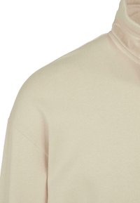 Maglione beige con collo alto che mostra dettagli sulle spalle e sul colletto, realizzato in un tessuto morbido e testurizzato con cuciture pulite su uno sfondo semplice.