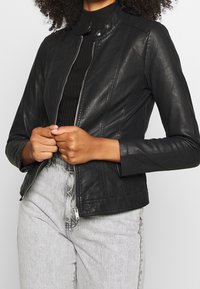 Black leather jacket with a stand-up collar, zip closure, and fitted design. Paired with high-waisted light gray jeans.