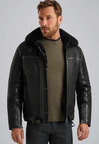 Man in zwarte shearling leren jack over olijfgroen gebreid sweater en donkere spijkerbroek tegen een grijze achtergrond.