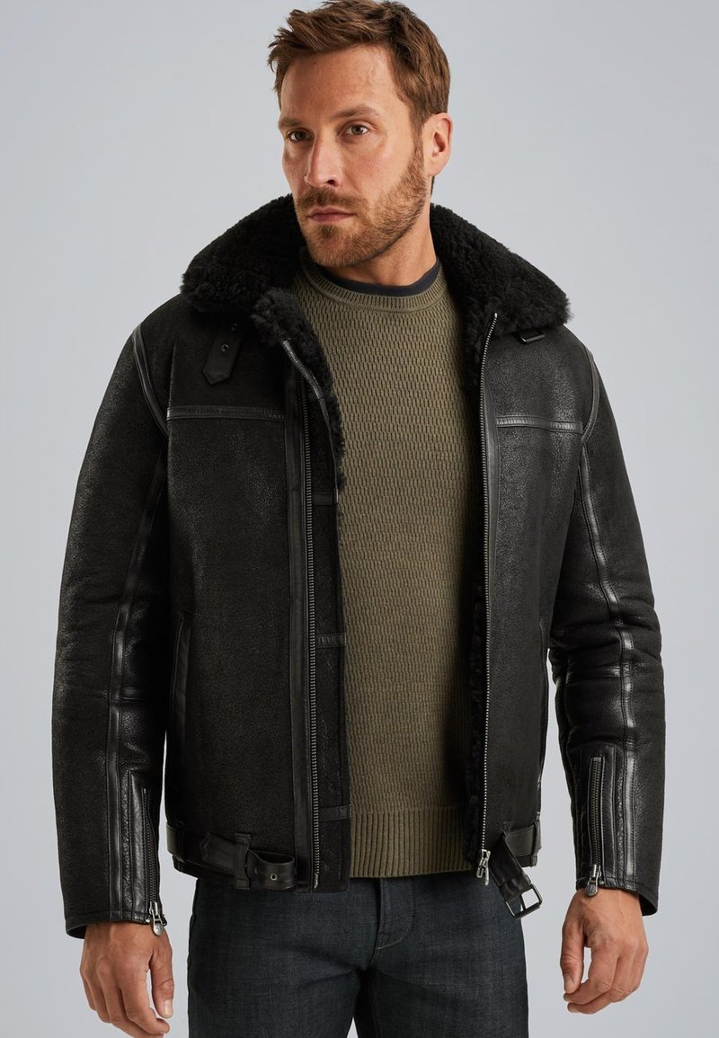 Man in zwarte shearling leren jack over olijfgroen gebreid sweater en donkere spijkerbroek tegen een grijze achtergrond.