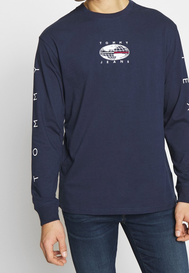 Marinblus med lång ärm som har en central Tommy Jeans-logotyp, med vit text på ärmarna och rund halsdesign. Mjuk bomullsmaterial.