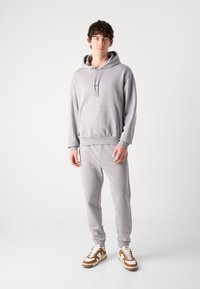 Pelēka kapuce ar kangaroo kabatu, saskaņotie pelēkie joggeri un baltas-brūnas sporta apavus. Fleece materiāls, brīvs piegriezums un regulējama kapuces aukla.