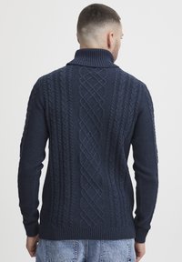 Pull en maille bleu marine avec un col montant, présentant un motif en relief de câble au dos et des poignets et ourlet côtelés.
