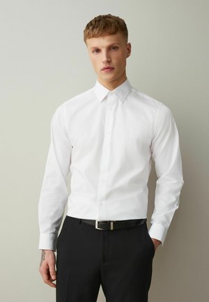 Young man with short hair wearing a white buttoned dress shirt tucked into black pants, standing against a plain light gray background.