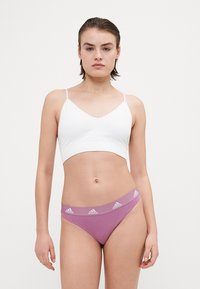 Bílý bezdrátový bralet s tenkými ramínky, spárovaný s fialovými tangy s logem na pásku v pase. Hladká látka, minimalistický design.
