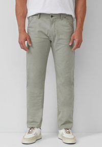Pantalon en coton vert clair avec une coupe slim, doté de poches avant et d'une fermeture éclair. Associé à des baskets blanches et un t-shirt blanc uni.