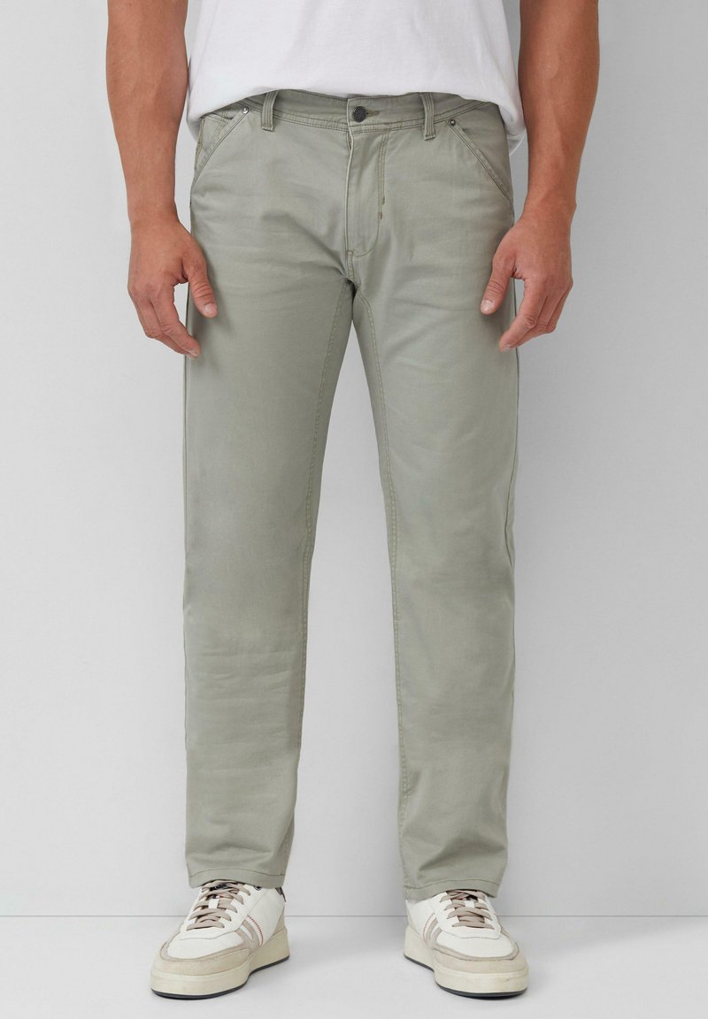 Pantalon en coton vert clair avec une coupe slim, doté de poches avant et d'une fermeture éclair. Associé à des baskets blanches et un t-shirt blanc uni.