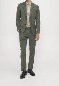 Uomo che indossa un completo slim-fit verde oliva con una camicia beige a bottoni e scarpe di pelle nere, in piedi con una mano in tasca.