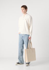 Ανοιχτό κρεμ πουλόβερ με γιακά, δύο κουμπιά, και σήμα λογότυπου. Συνδυάζεται με ανοιχτό μπλε τζιν και καφέ καμβά τσάντα φόρτωσης. Υποδήματα είναι μαύρα αθλητικά παπούτσια.