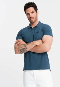 Tmavě modrá polo tričko vyrobené z bavlny, s límečkem, trojím knoflíkovým zapínáním a logem na hrudi. Hladká textura, přiléhavý střih.
