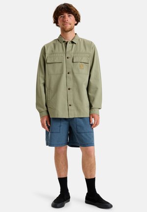 Olivovo zelená košeľa na gombíky s dvoma prednými vreckami a logom, spárovaná s modrými džínsovými šortkami a čiernymi slip-on topánkami.