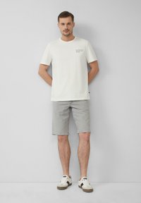 T-shirt blanc en coton à manches courtes, avec un texte sur la poitrine gauche, associé à un short en denim gris et des baskets blanches avec des accents marron.