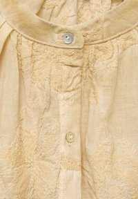 Camicia di lino color crema con delicata ricamo floreale, colletto arrotondato e chiusura con un solo bottone. Texture morbida, dettaglio sottile.