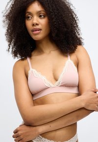 Naisel kihara juustega, seljas heleroosa pitsiga kaunistatud rinnahoidja ja sellele sobivad pitsist aluspüksid, käed risti üle keha.