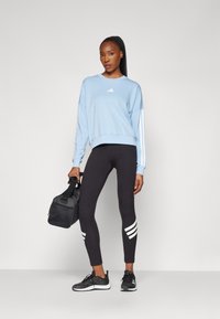Lyseblå hettegenser med hvit Adidas-logo, kombinert med svarte leggings med hvite striper ved leggen. Svart treningsbag og atletiske sko.
