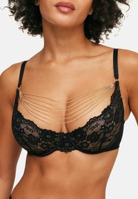 Soutien-gorge en dentelle noire avec une conception de coupe structurée, présentant des accents en chaîne dorée tombant sur le décolleté. Les bretelles ajustables complètent le look.