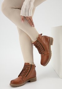 Stivaletti in pelle marrone con cerniera laterale, punta tonda, lacci anteriori e suola in gomma testurizzata. Indossati con leggings a coste beige.