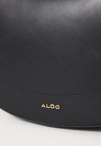 Černá kožená kabelka s hladkou texturou, zakřiveným designem a zlatým detailem loga "ALDO" blízko spodní části. Obsahuje prošívané švy.