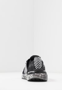 Zapatilla deportiva negra con una suela de diseño en gris y negro, cuerpo de malla texturizada y acentos con rayas en el talón. Diseñada para ofrecer soporte y flexibilidad.