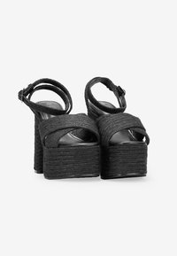 Sandalias de plataforma negras con correas de tela trenzada, diseño de punta abierta y cierre de hebilla en el tobillo. Suelas gruesas y texturizadas para mayor altura.