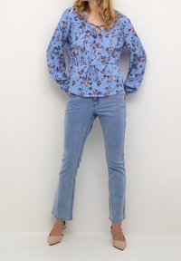Blusa floreale azzurro con maniche lunghe, vita regolabile con cordoncino e scollo rotondo abbinata a jeans azzurri a campana e tacchi alti beige.
