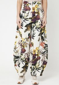 Vrouw in losse, hoge taille broek met een meerkleurige bloemenprint, gecombineerd met witte sneakers, staande tegen een effen achtergrond.