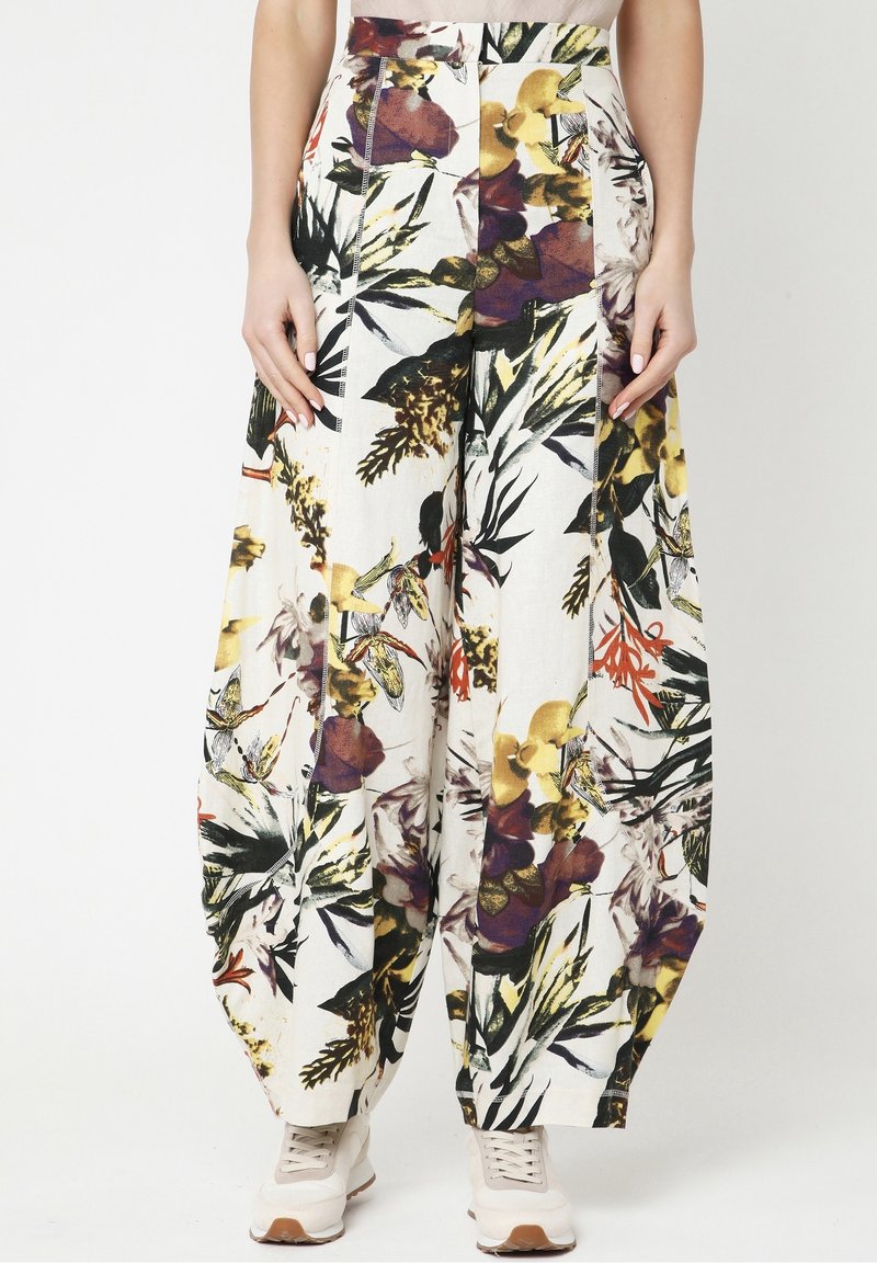 Vrouw in losse, hoge taille broek met een meerkleurige bloemenprint, gecombineerd met witte sneakers, staande tegen een effen achtergrond.