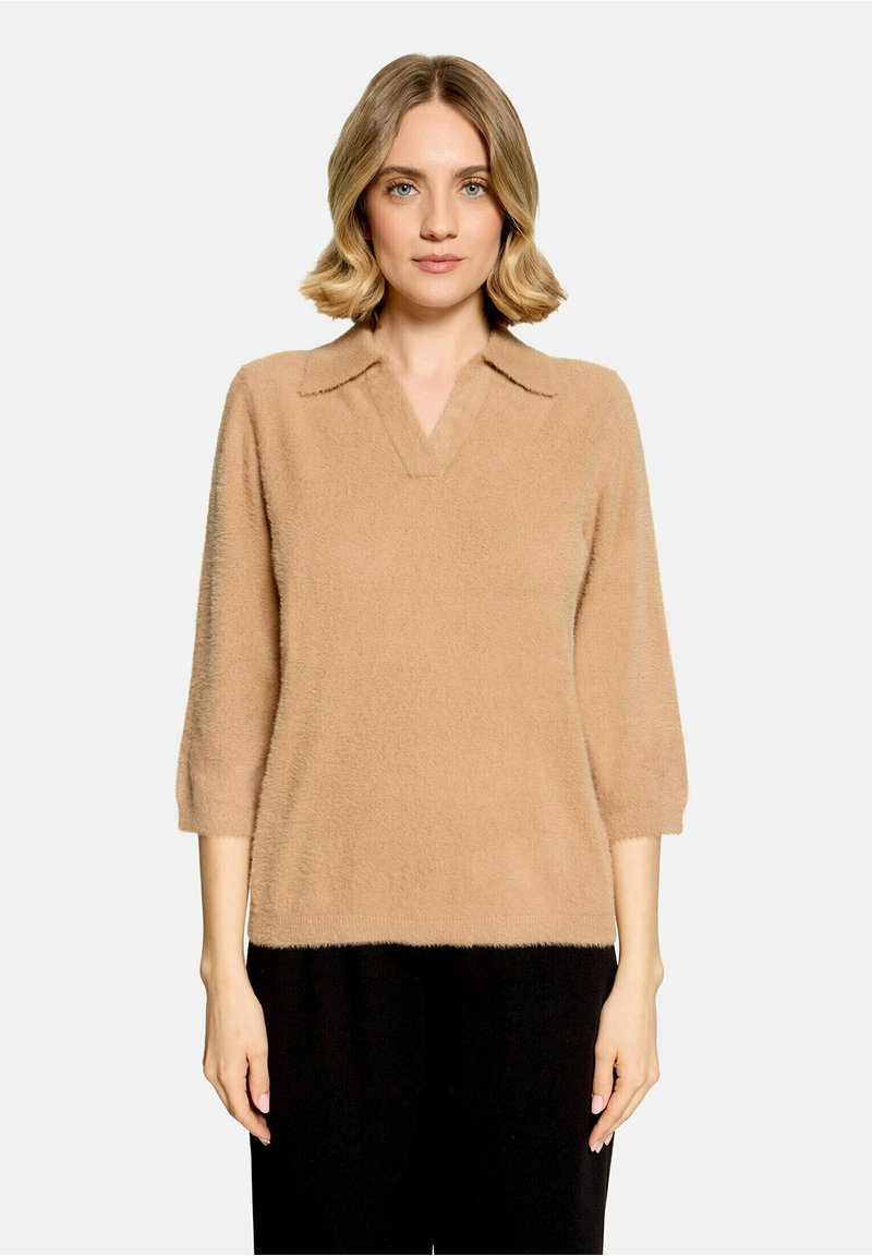 Femme aux cheveux blonds courts portant un pull beige duveteux avec un col et des manches trois-quarts, debout devant un fond clair uni.