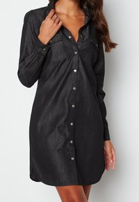 Svart denimklänning med långa ärmar, två bröstfickor och silverknäppningar. Har en rak skärning och sidoflikar för ökad rörlighet.