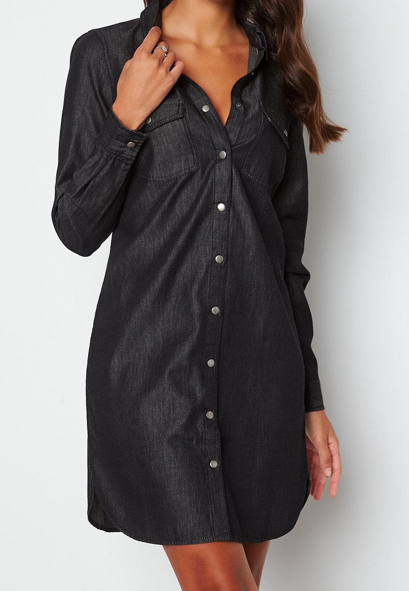 Svart denimklänning med långa ärmar, två bröstfickor och silverknäppningar. Har en rak skärning och sidoflikar för ökad rörlighet.