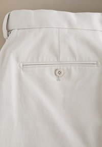 Pantalon en tissu beige avec une poche arrière passepoilée sécurisée par un bouton à quatre trous assorti et des passants de ceinture à la taille.