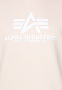 Sudadera de algodón beige con un gran logo blanco que incluye el texto "Alpha Industries" y "Knoxville, Tennessee, U.S.A." Textura suave.