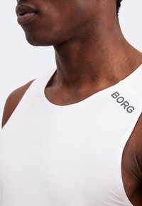 Κοντινό πλάνο ενός ατόμου που φοράει λευκό αμάνικο τοπ με τη λέξη "BORG" τυπωμένη κοντά στον δεξί ώμο.